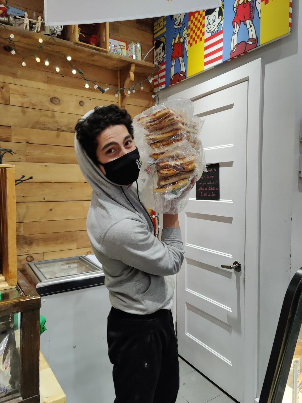 A masked volunteer in a grey hoodie holds up a large bag of wrapped sandwiches in a small kitchen.