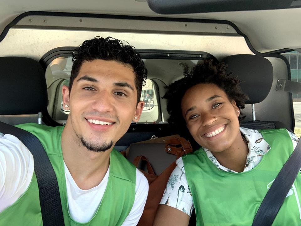 Two smiling volunteers in green vests take a selfie in a car, seatbelts on.