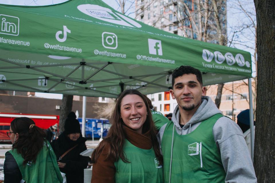 Two volunteers in green vests smile side by side under the green Welfare Avenue tent.