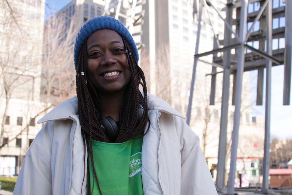 A smiling volunteer in a blue beanie and white coat over a green Welfare Avenue vest, downtown.