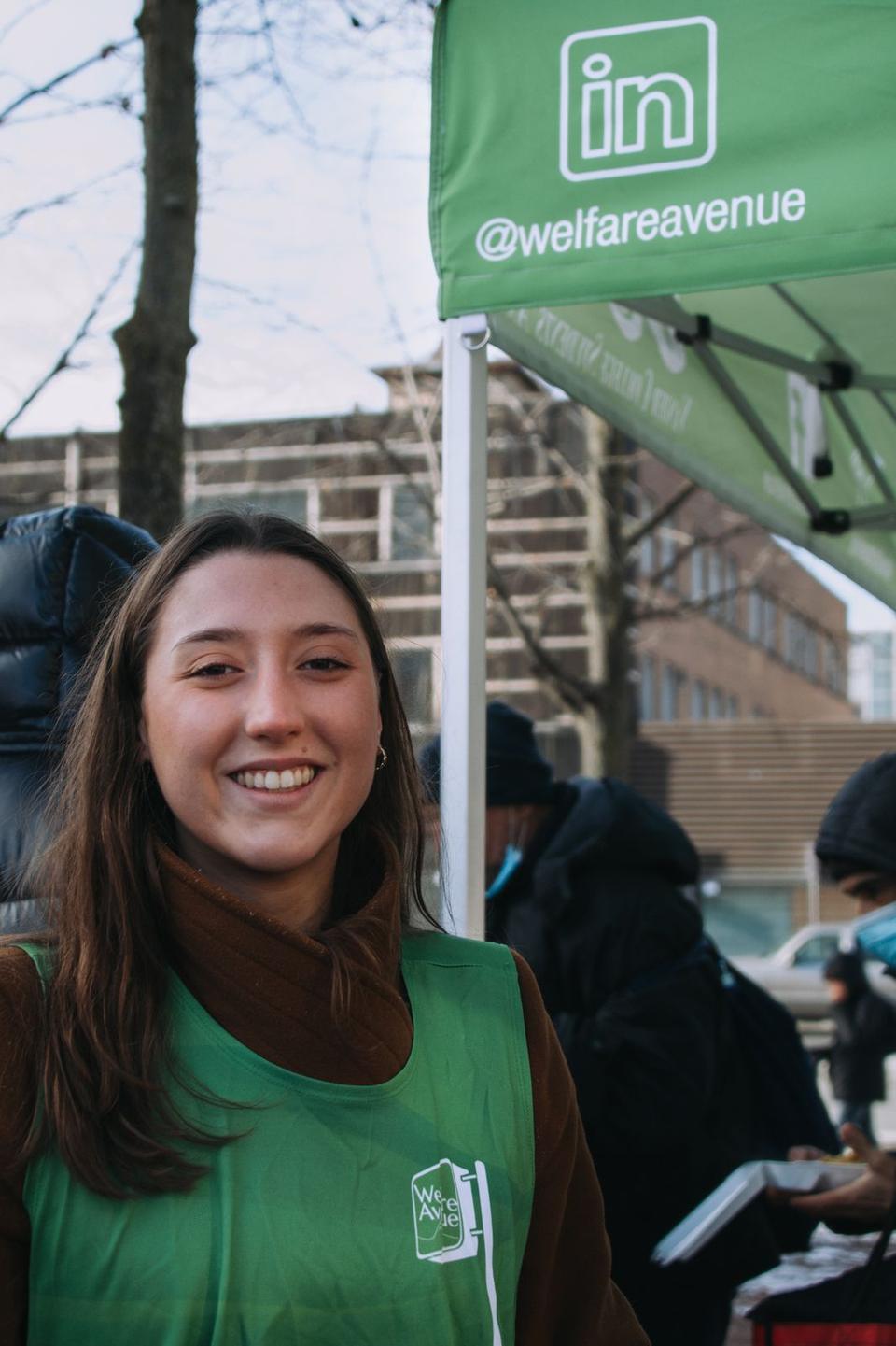 A smiling volunteer in a green vest over a brown jacket stands under the tent flap printed with the @welfareavenue handle.