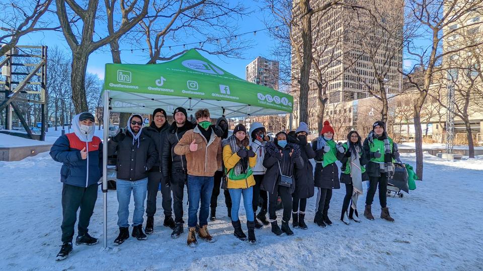 About thirteen volunteers in winter coats and green vests pose in the snow in front of the green tent under bare trees.