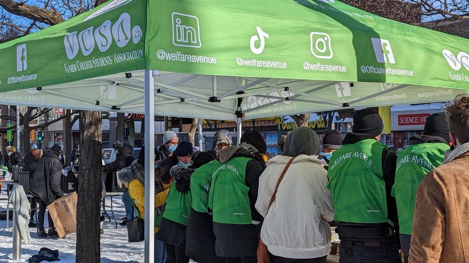 Volunteers in green vests, seen from behind, serve under the Welfare Avenue tent in winter.