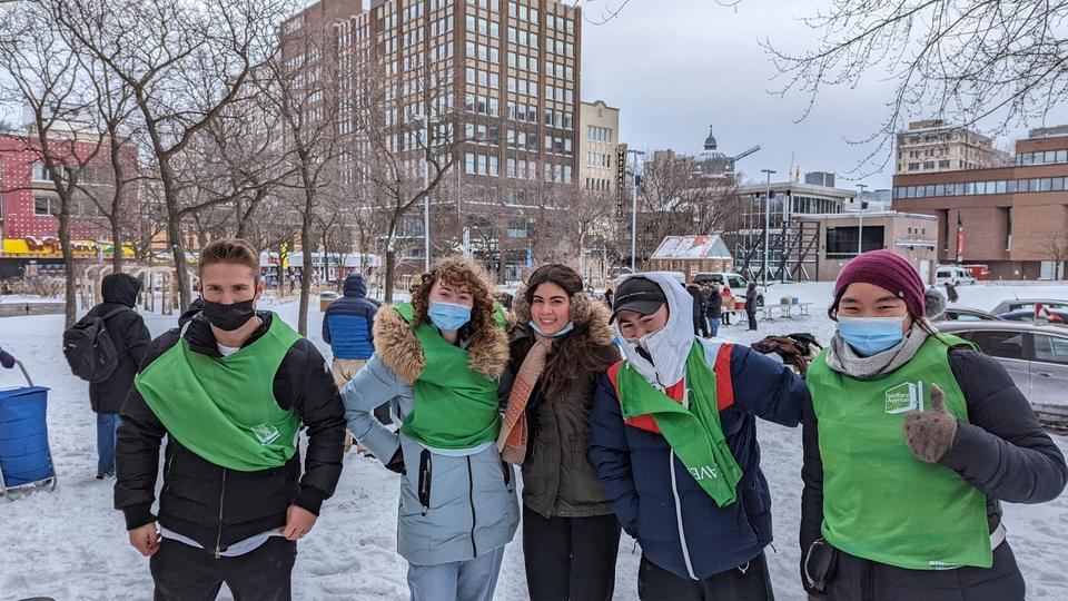 Five volunteers in winter coats and green Welfare Avenue vests pose in the snow, one giving a thumbs up.