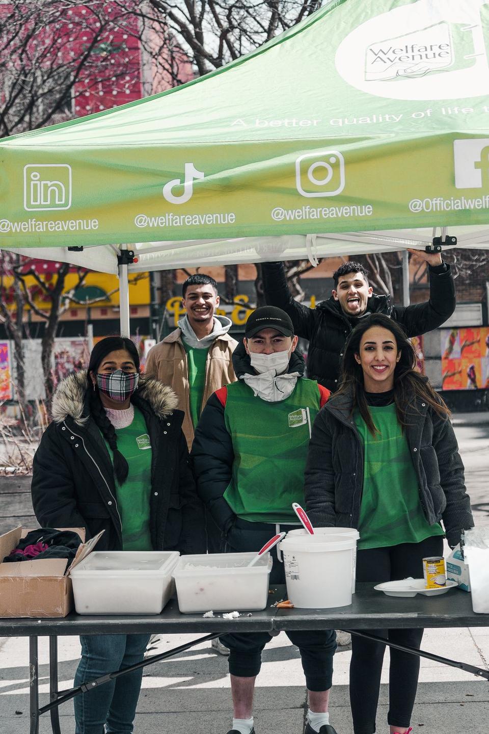 Five volunteers in green vests, some masked, pose behind the serving table under the tent.