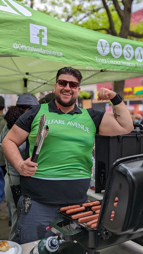 A volunteer in sunglasses and a Welfare Avenue vest flexes his arm with tongs in hand beside a grill of hot dogs.