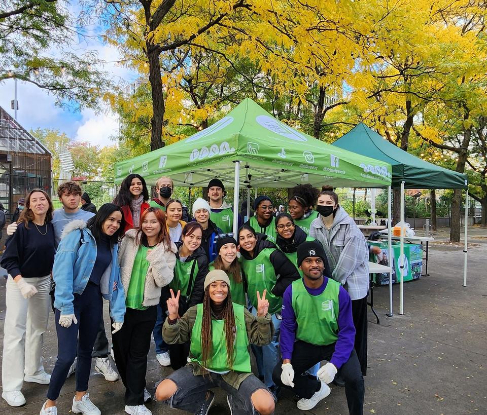 About twenty volunteers, many in green Welfare Avenue vests, pose in front of two green tents under yellow autumn trees.