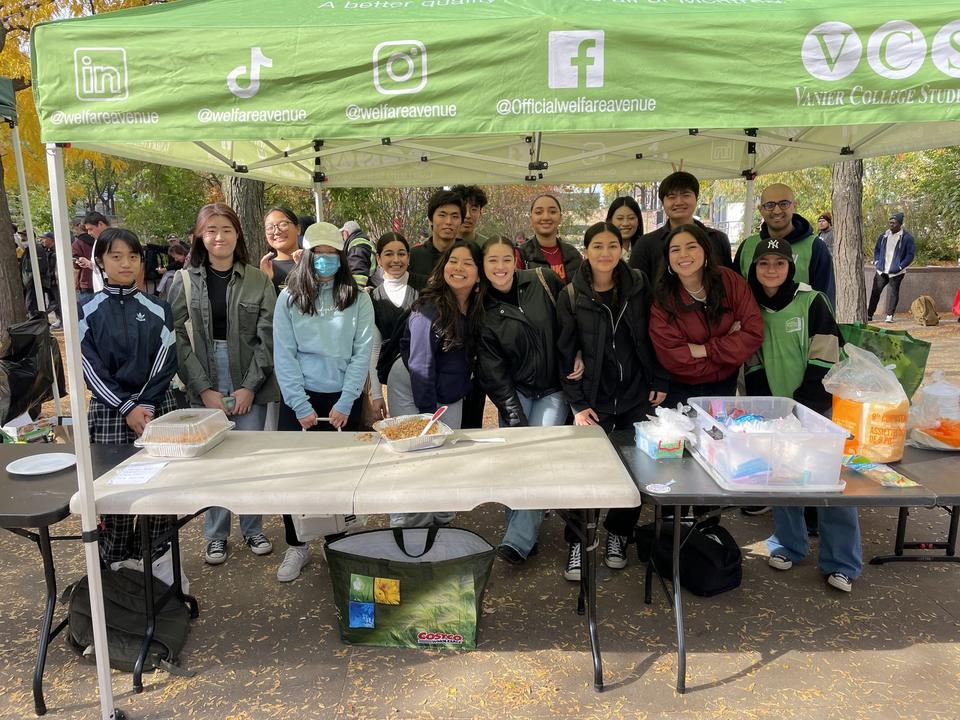 About twenty-five young volunteers stand smiling behind folding tables under the green Welfare Avenue tent.