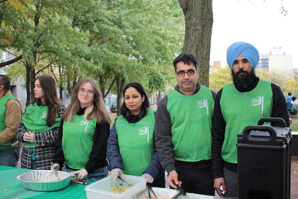 Six volunteers in green vests smile behind the serving table in a park.