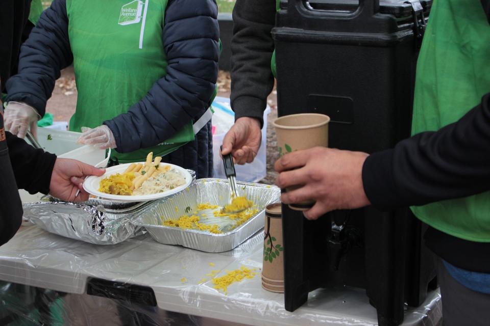 Volunteers in green vests serve food onto a plate held out across the table, beside a drink dispenser.