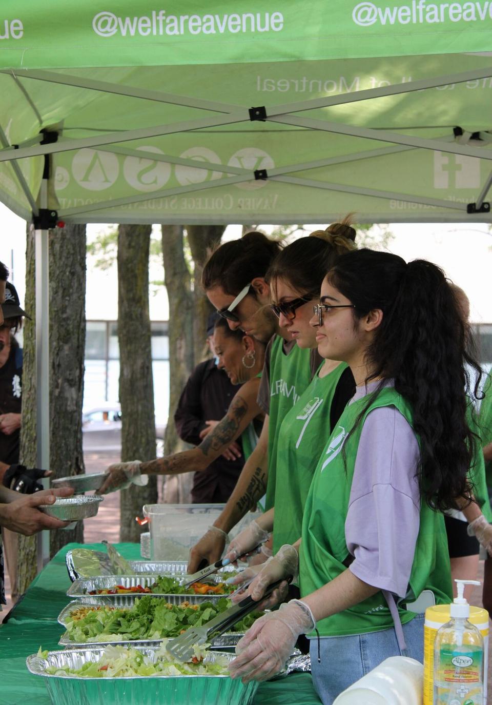 Four volunteers in green vests and gloves serve salad from foil trays along the table under the tent.