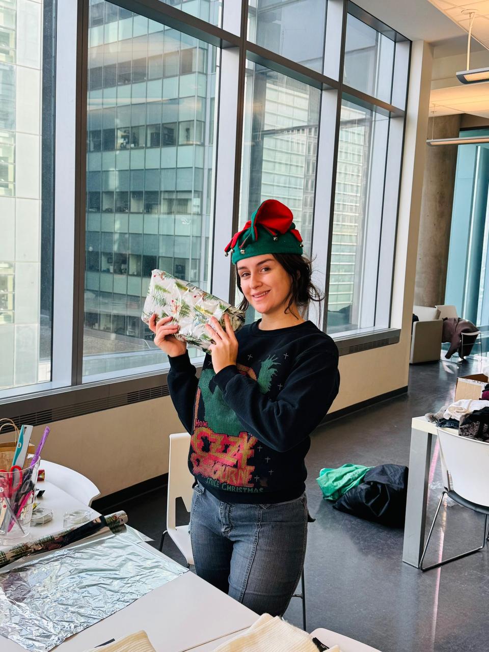 A smiling volunteer in a green elf hat holds up a wrapped gift.
