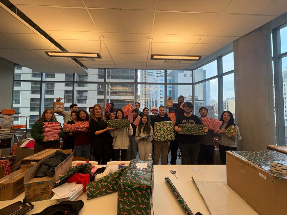 About twenty volunteers hold up wrapped gifts behind tables in a bright room with city windows.