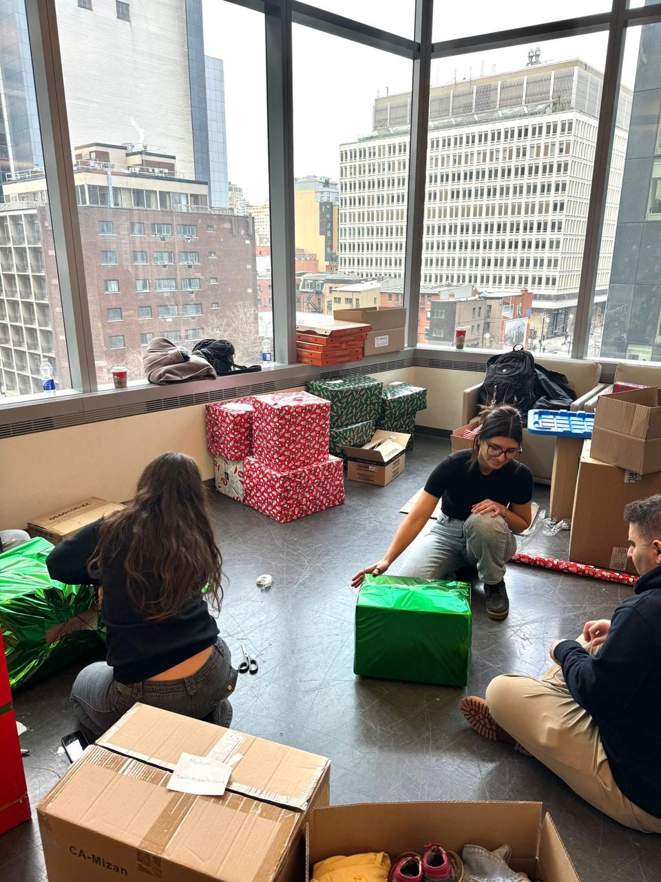 Volunteers sit on the floor wrapping gifts beside large windows and stacks of boxes.