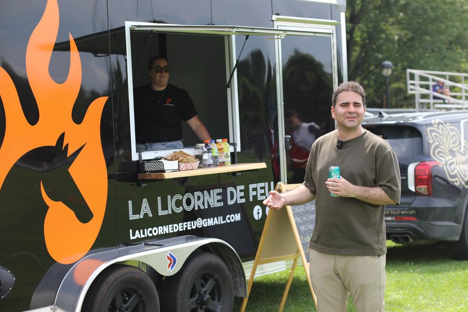 A smiling man holding a can stands beside the black La Licorne de Feu food trailer, a cook at its window.