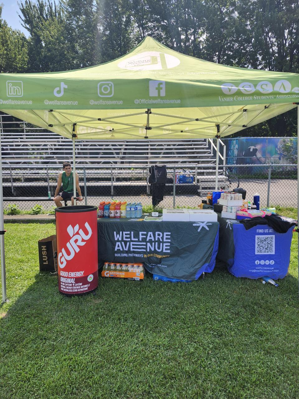 Our green tent with the Welfare Avenue table banner and a red GURU drinks bin, bleachers behind.