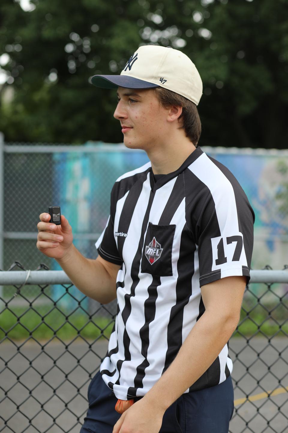 A referee in a striped shirt and a cream cap stands by the field.