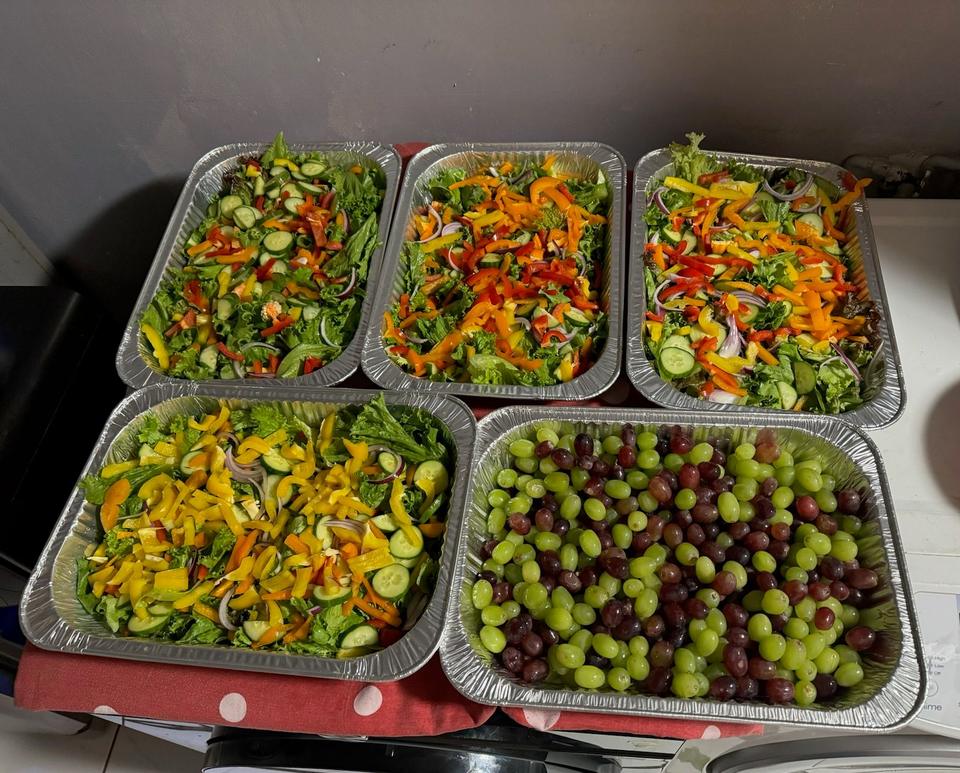 Foil trays of green salads with cucumber and red onion, and a tray of grapes, seen from above.